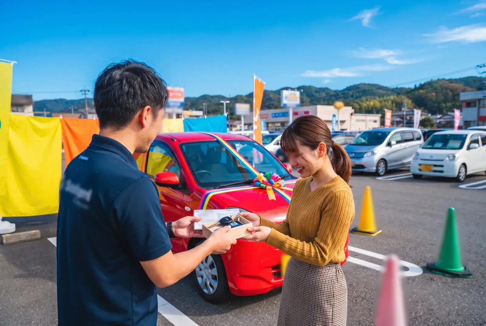 のぼり旗の並ぶ展示場で、お客様に車の鍵を手渡すスタッフ
